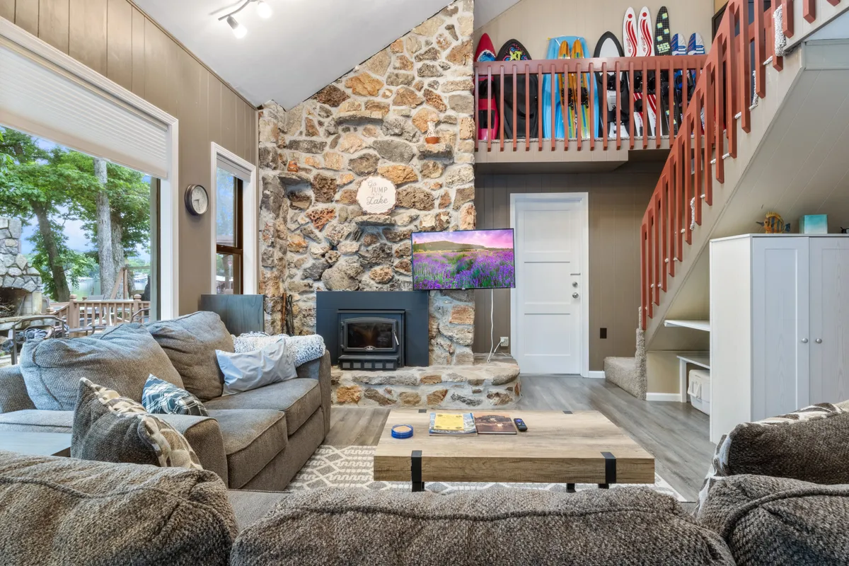 A bright, inviting living room featuring a prominent stone fireplace, large lake-facing windows that flood the space with natural light, a cozy seating area, a wooden staircase, and colorful surfboards propped nearby. The scene conveys an atmosphere of relaxation and togetherness, enhanced by the views of nature outside.