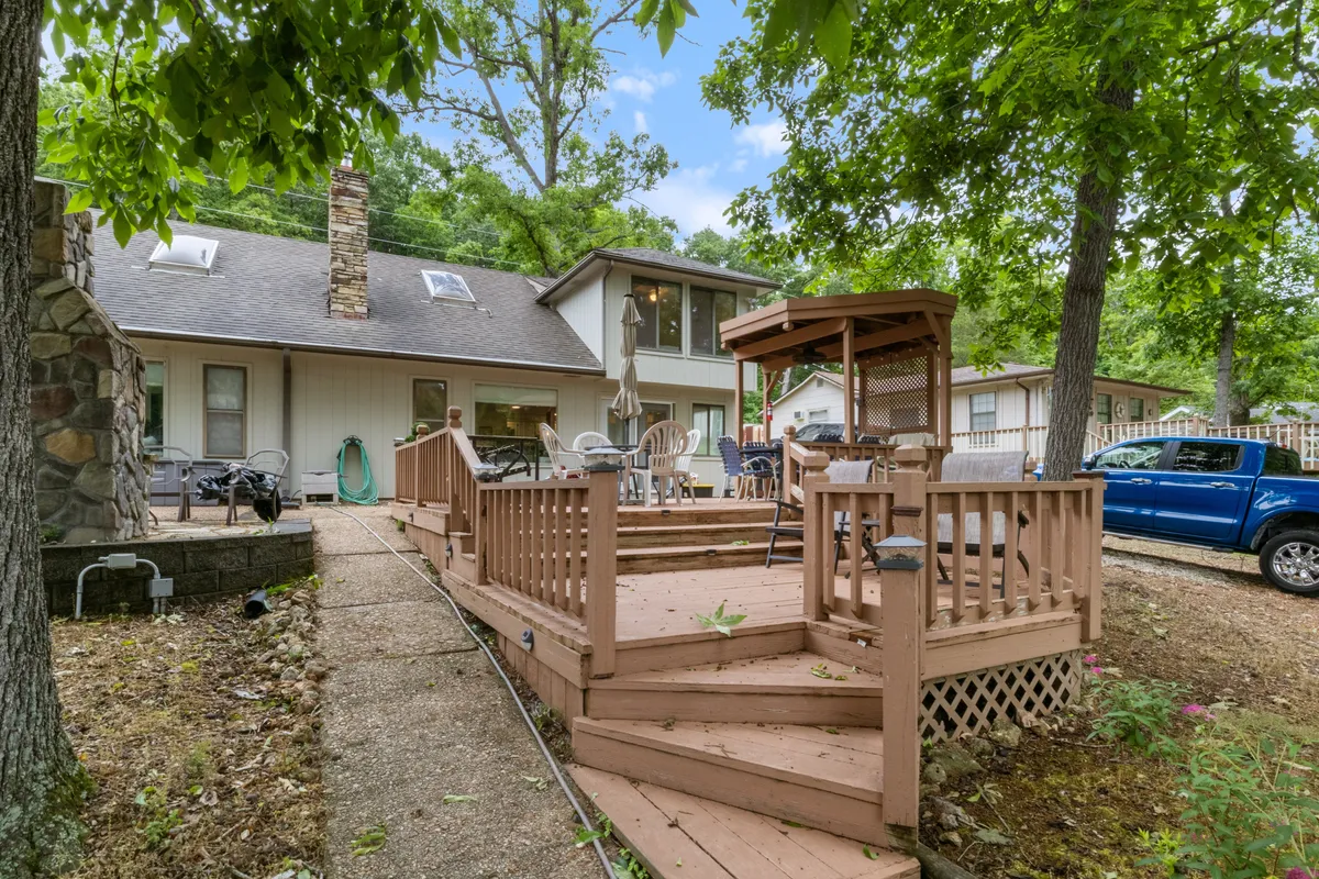 A spacious outdoor deck featuring a gazebo, wooden railing, and a prominent stone chimney. The seating area invites outdoor dining or relaxing, surrounded by lush greenery and set against a tranquil lakeside backdrop.