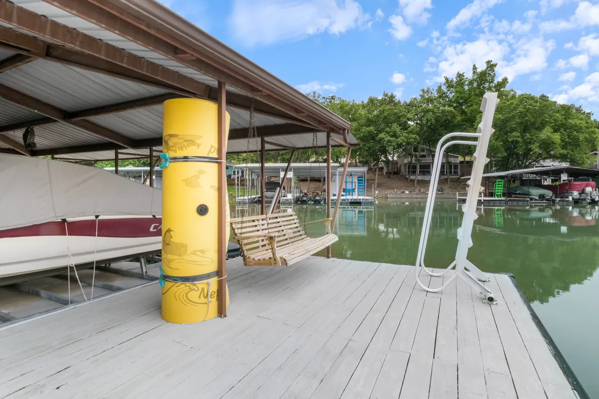 A lively lakeside scene with a yellow buoy, a swing, dock, and a moored boat. The area is surrounded by greenery and offers space for fishing, relaxing, or taking in the water views, evoking a sense of leisure and outdoor enjoyment.