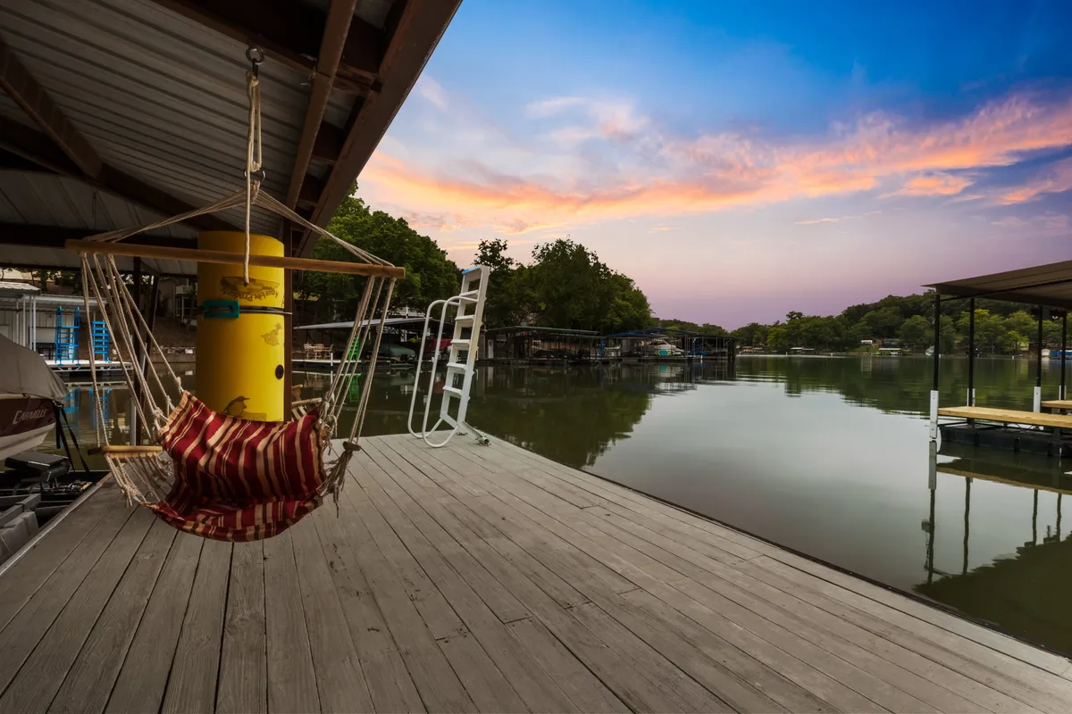 The image showcases a serene lakefront setting with a hammock gently strung between posts, inviting guests to relax while enjoying sweeping views of the tranquil water. The private dock extends into the quiet cove, perfect for fishing or launching a kayak. Sunlight glistens on the lake nearby, and there's ample space for lakeside lounging and barbecues.