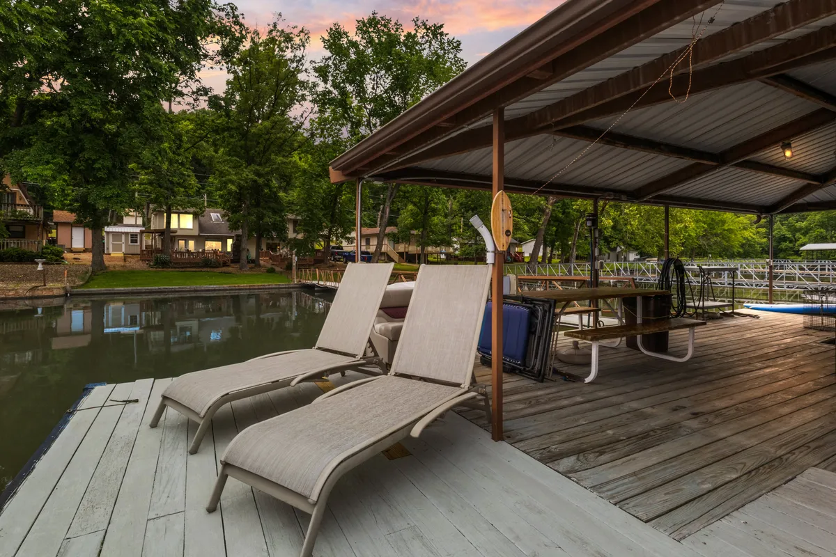 This exterior shot captures a serene lakeside scene with lounge chairs set up on a dock, surrounded by trees and offering views of the water and neighboring houses. The setting suggests a perfect spot for relaxing, fishing, or watching kids swim and play by the water's edge.