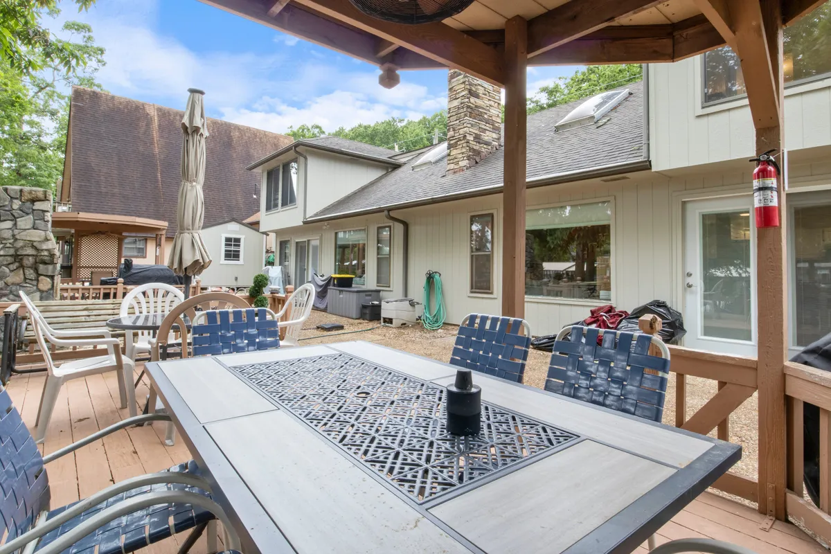 An outdoor dining table sits on a patio adjacent to a stone chimney, surrounded by greenery. The arrangement invites gatherings for meals or sunset watching outdoors, with patio seating offering a view over the lakeside landscape.