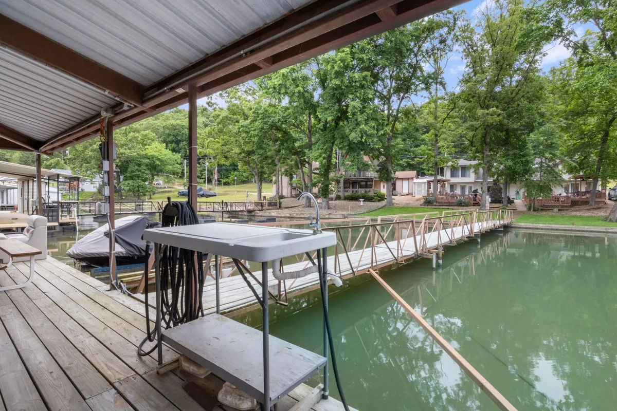 The image shows a dock reaching out over the water, with moored boats and trees providing a scenic lakeside backdrop. The space invites groups to gather for fishing, swimming, or just hanging out together by the water.