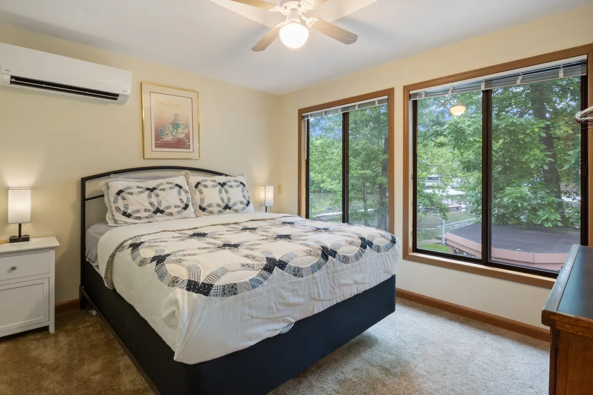 A tranquil bedroom featuring a comfortable bed flanked by nightstands, with large windows letting in natural light and a ceiling fan overhead. The space is accented by tasteful wall art and offers a view of the outdoors, reinforcing a restful retreat atmosphere.