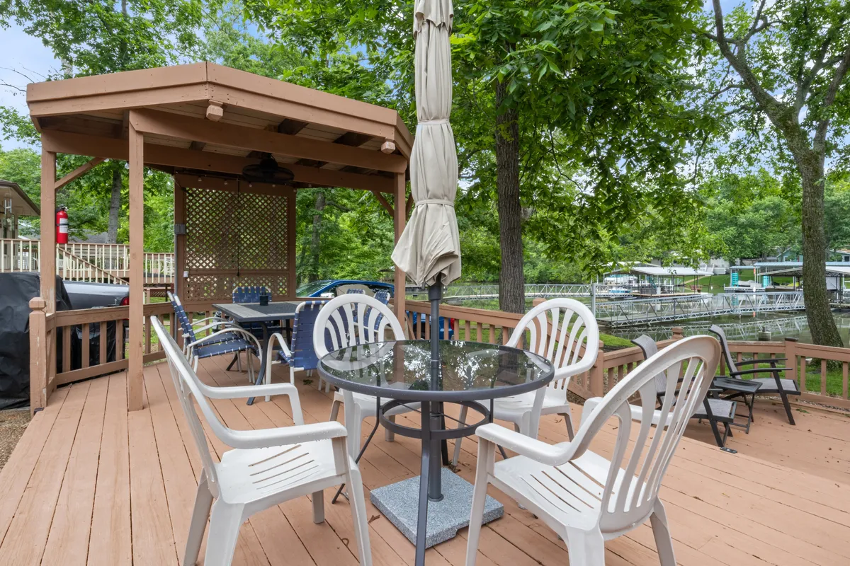 This image features an outdoor dining table beneath a pergola, surrounded by lush greenery with a boating dock visible in the background. The setting is ideal for social gatherings, dining, and relaxing by the water, all while soaking in the tranquility and natural beauty of the lakefront.