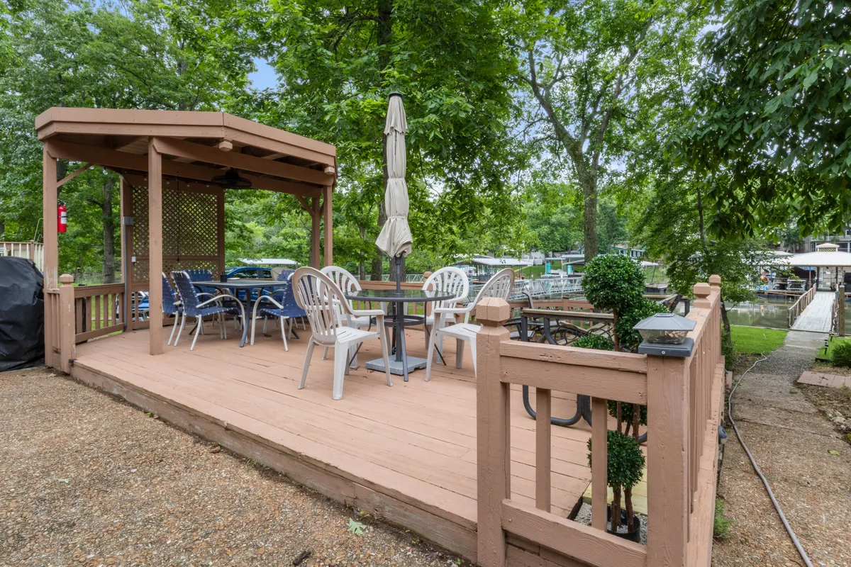 This image shows a wooden deck equipped with an outdoor dining table shaded by umbrellas, surrounded by lush greenery. The setting evokes the idea of casual family meals or barbecue evenings just steps from the lake.