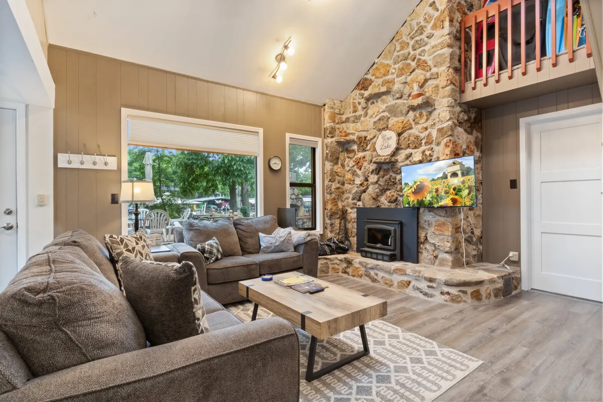 A warm, welcoming living space featuring a stone fireplace, comfortable seating arranged for conversation, a wooden coffee table, and a large window that allows in natural light and offers a view of the outdoors. The overall feel is both cozy and airy, suitable for gatherings or quiet moments by the fire.